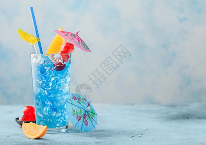 高球玻璃中的蓝色环礁湖夏季鸡尾酒配有甜樱桃和橙色切片蓝桌底有雨伞图片