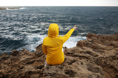 身穿黄色雨衣的年轻女士坐在悬崖上看着海浪的巨同时在雨天岩滩的阴云春季气中享受美丽的海景身穿黄色雨衣的年轻女士坐在悬崖上看着海浪的图片