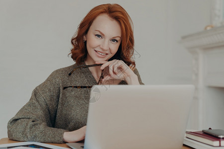 红发职业女商人在笔记本电脑上远程工作阅读网上商业事务的文章坐在桌面上使用现代技术编写财务报告图片