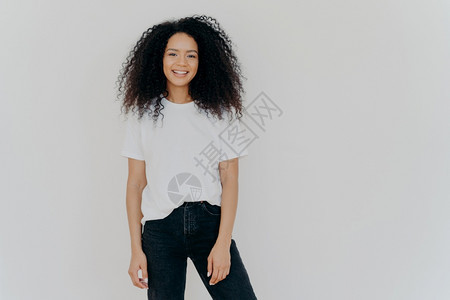 相片中微笑着的女士卷黑头发身材瘦弱穿着白色T恤衫和黑牛仔裤面容表现愉快白色背景模特复制广告内容的空间图片