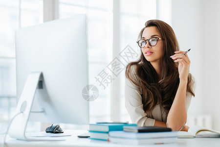 戴眼镜的女人黑发女士专注在大监视器上坐在桌面拿着笔写记戴眼镜进行视力矫正在工作空间装扮背景