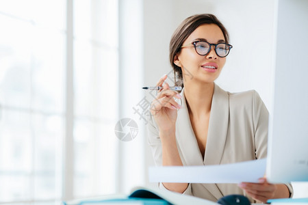 繁荣的女商人关注计算机监测研究金融问题持有证件和笔在办公室工作穿正规服装和眼镜穿白色背景的服装和眼镜背景图片