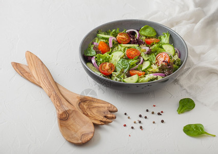 新鲜健康的素食蔬菜沙拉配有生和西红柿洋葱和菠菜放在白桌底的灰色碗盘中配有叉子和勺图片