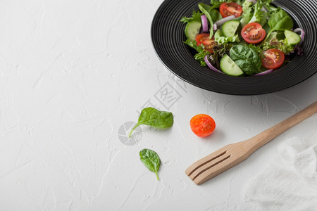 新鲜的健康素食蔬菜沙拉配有生和西红柿洋葱和白底黑碗的菠菜配有叉和白厨房布图片