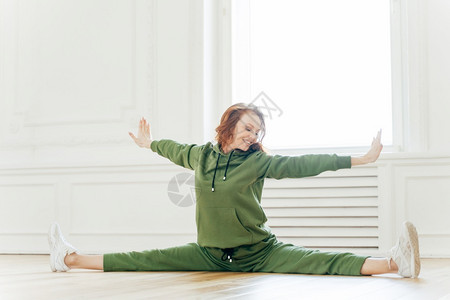 身体健康的女室内镜头伸展手和腿的侧面坐成裂缝的姿势向右倾斜的头部牙齿笑容红发穿运动服在工作室的姿势从事柔软的瑜伽图片
