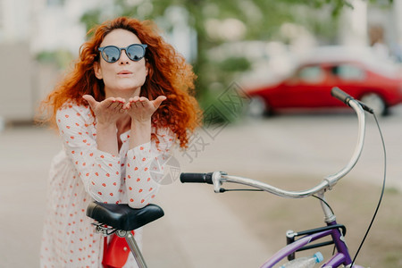 深爱的红发妇女向街上情人发出空吻将棕榈贴在嘴边穿圆窗帘衣服骑自行车在户外积极休息在街上摆姿势娱乐时间概念图片
