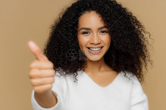 黑暗皮肤健康卷发年轻女人的头部在镜上露出拇指笑得愉快给予认可或喜欢的想法穿白色跳跃器孤立在棕色工作室背景上图片