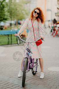 每周在城市骑自行车的卷发红妇女被垂直射杀有闲暇时间穿长裙遮光和运动鞋携带小红袋手持渔具图片