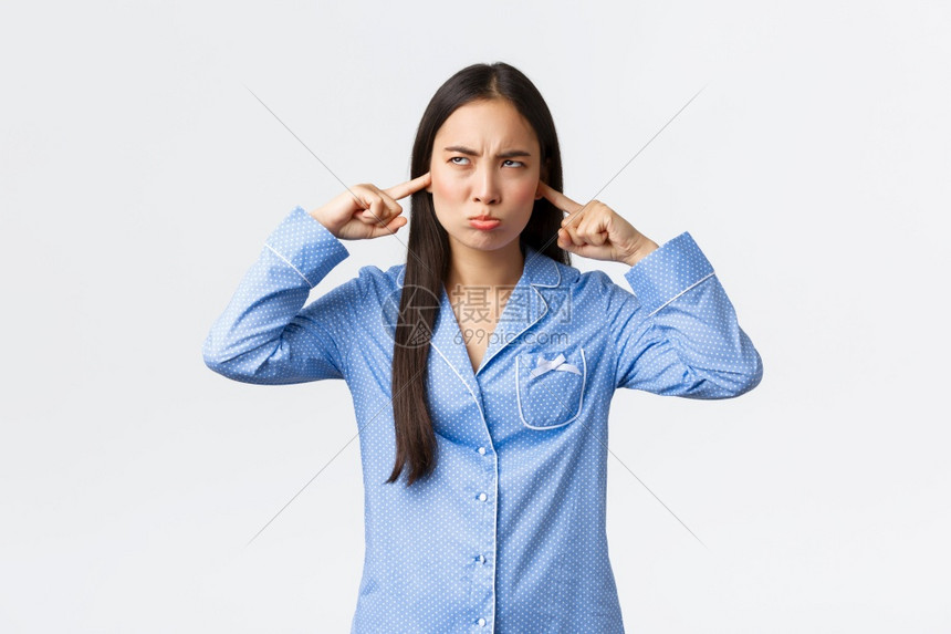 愤怒的亚洲女孩穿着蓝睡衣抱怨大学宿舍里的邻居大声看着烦恼和不高兴的邻居看着吵闹和不高兴的邻居闭上耳朵远离可怕的噪音站着白色背景愤图片