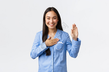 身穿蓝睡衣的诚实真和的亚洲女孩举起一只手将握在心上如许下诺言说出真相或发誓微笑如在白色背景上戴着果酱身穿蓝色睡衣举起一只手将握在背景图片