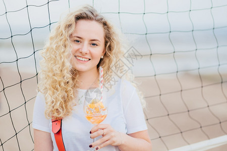 可爱的欧洲年轻快乐女人长着卷毛轻笑拿着橙子鸡尾酒杯站在网球附近图片