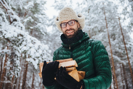 长着胡子的男戴着眼罩和带动脉棒的温暖帽子拿着木柴用白雪扑向树木在森林中度过时间快乐男人的户外镜头躺在森林中图片