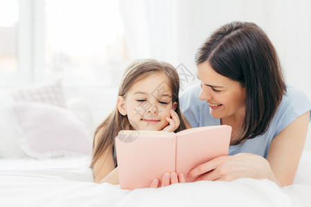带着深色头发的年轻母亲快乐地看着她的小女儿一起读有趣的书或童话故事在家里享受居用餐人庭童年概念图片