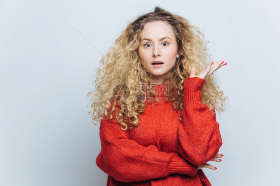 美丽的蓝眼睛金发美女看着镜头的迷惑穿着松散的红毛衣不满什么孤立在白背景上令人惊讶的概念图片