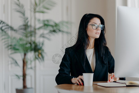 集中的女企业家穿着优雅的黑色服装在计算机工作坐在舒适场所的桌子上穿着透明的眼镜浏览互联网使商业项目考虑战略背景图片
