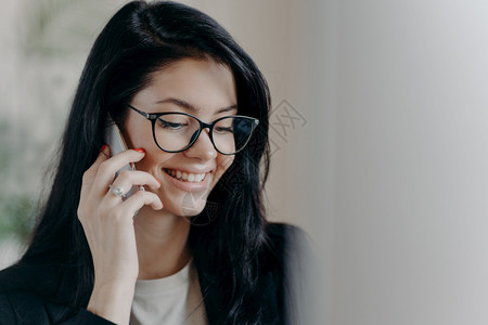 快乐的女律师将手机放在近耳旁与客户愉快地交谈讨论工作议题笑容温柔戴透明眼镜正式穿戴图片
