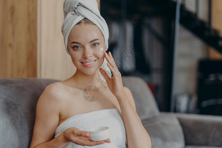 有健康光滑皮肤的漂亮年轻女子施用面霜淋浴后头部涂上毛巾在舒适沙发上涂毛巾卫生程序美容和概念图片