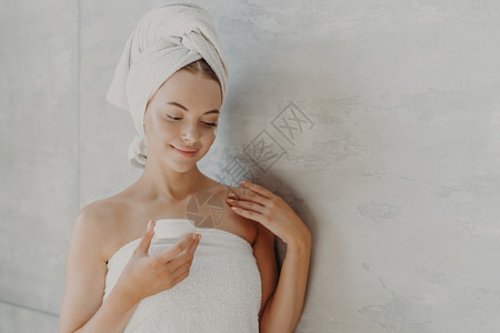 穿着健康皮肤的美容女人最起码的化妆品涂面霜淋浴后被裹在巾上灰色背景附近图片