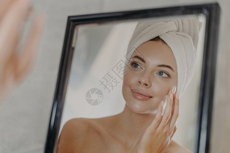 欧洲年轻女在脸上触摸柔软的皮肤在浴室镜子中看着自己洗完澡后头戴包着白毛巾肩膀脱下图片