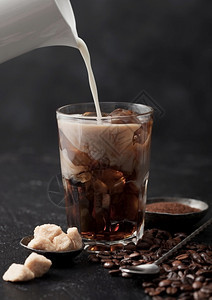将新鲜牛奶倒入玻璃加冰咖啡豆和甘蔗糖地面咖啡和黑桌底的长勺子图片