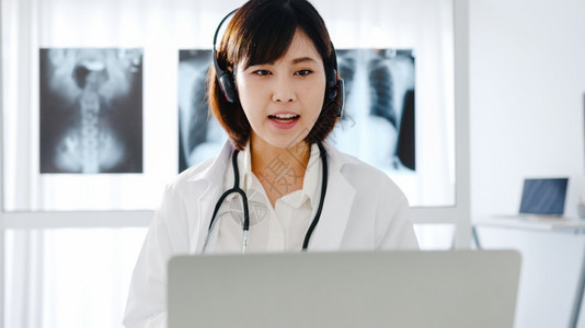 身穿白色医疗制服有听诊器的年轻女医生使用电脑笔记本在医疗诊所或院的服务台与病人交谈视频电话图片