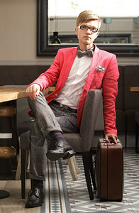 年轻帅的时装模特身着眼镜的年轻时装帅男时装模特身着明亮红色夹克领结带和手提箱在室内等候图片