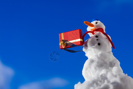 穿着蓝色螺丝顶上的小快乐雪人戴着帽子红围巾外出有叉子圣诞礼物盒冬季节特制蓝天空背景图片