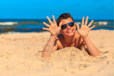 13岁快乐的笑男孩在海边展示了沙中的双手浅田地深度专注在手和脸上图片