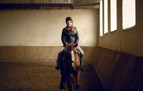 坐在室内竞技场的棕马上年轻女骑手的肖像图片