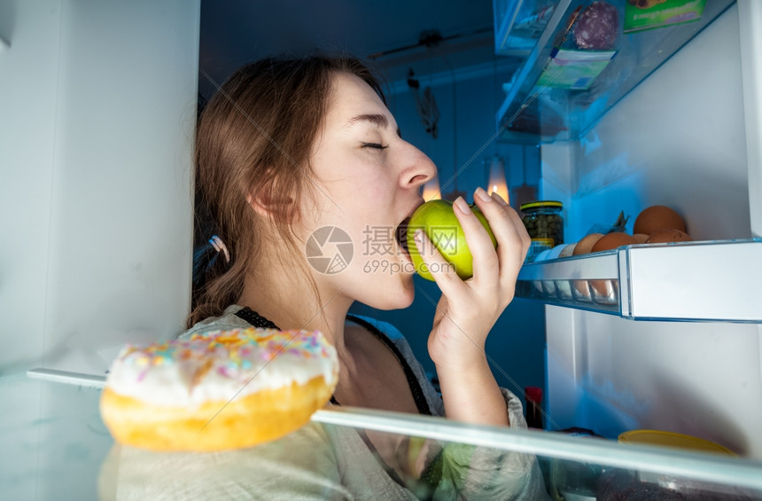 坐在冰箱里看着吃绿苹果的年轻女人近距离肖像图片