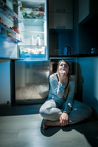 身穿睡衣的妇女晚上在冰箱旁边的地板上吃东西照片图片