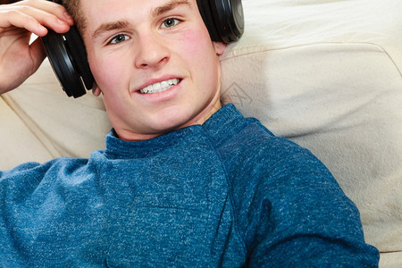 英俊的年轻人带着耳机躺在沙发上家里听音乐图片