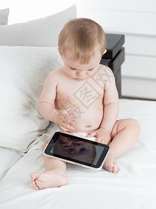 穿着尿布坐在床上玩数字平板电脑的可爱婴儿男孩图片
