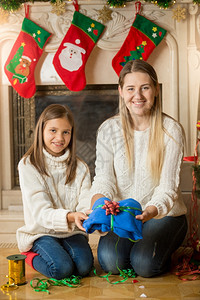快乐的年轻母亲和女儿坐在壁炉的地板上为圣诞礼物打包毛衣图片