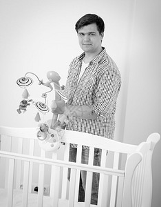 年轻有爱心的父亲黑色和白形象将婴儿和s床和将玩具旋转木马放入其中图片