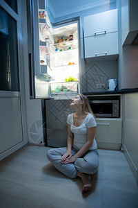穿着睡衣坐在厨房地板上看露天冰箱的年轻美女图片