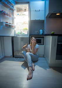 深色地板坐在厨房地板旁边的女士打开冰箱和吃披萨背景