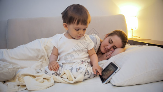 玩ipad的小孩小可爱男孩在母亲睡床上时使用数字平板电脑小男孩在母亲睡床上时使用数字平板电脑背景