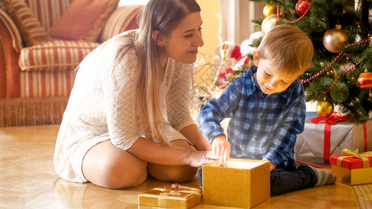 托德勒男孩与母亲坐在圣诞树下打开神奇发光的礼物盒小孩男与母亲坐在圣诞树下打开神奇发光的礼物盒图片
