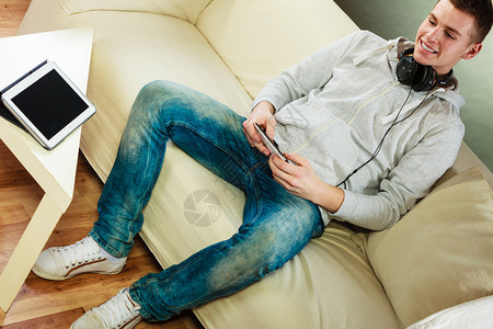 年轻人在沙发上休息时用耳机智能手和家用平板电脑图片