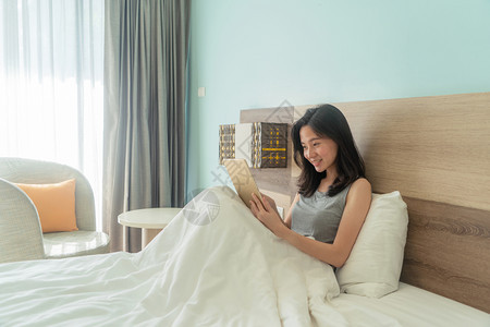 快乐的亚洲女人笑着用平板在床上现代卧室里早上带着白毯子图片