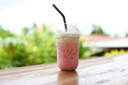 夏季在塑料杯果汁中喝粉红色草莓牛奶冰饮料在有自然背景的木制桌上吃草图片