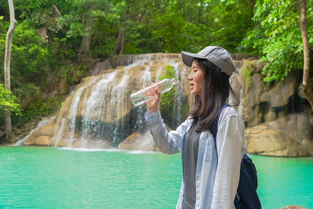 亚洲快乐妇女从热带森林瀑布的瓶子饮用水中在泰公园旅行和度假期间有树木图片