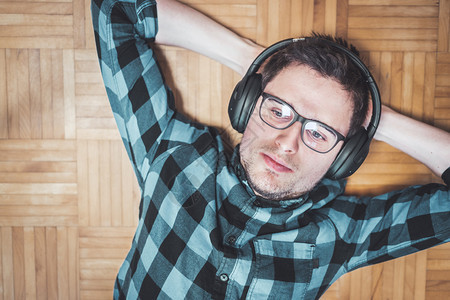 穿蓝衬衫的白人青年男子躺在木地板上用无线耳机录制音乐图片