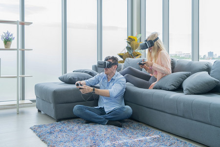穿着VR或虚拟现实眼镜的西方年轻夫妇坐在沙发上玩电子游戏的耳机当家门电视面前播放技术和创新概念玩得开心背景图片