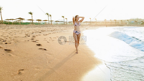 在沙海滨与日落的天空对望着日落的天空看着镜头在沙海滨与日落的天空对望着阳光的沙滩上长着发的美丽微笑着年轻女子肖像图片