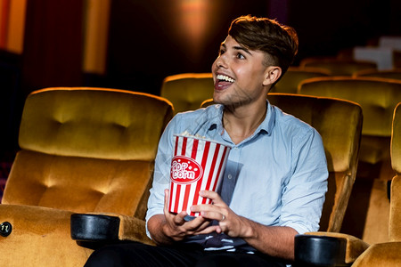 一个年轻男人笑着享受爆米花一边在电影院看图片