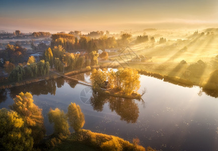 湖边小岛上有丰富多彩的树木房屋空中景象秋天是福吉的日出美丽景色与雾中村庄金色的太阳光束水中的反射在乌克兰倒塌图片