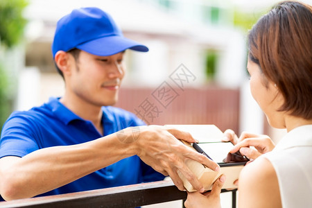 亚洲妇女在收到穿蓝衬衫男送货员的包裹后在便携式移动设备上签署电子名包件购物和数字标志概念图片