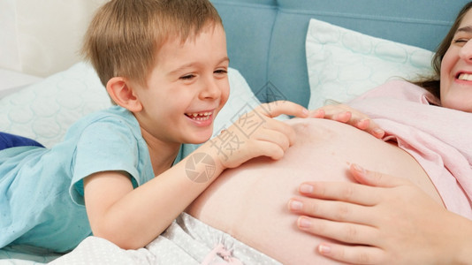 微笑的小男孩在床上打探和触摸着躺在床上的怀孕母亲大肚子怀孕准备和有孩子的概念微笑小男孩在床上打探和摸着怀孕母亲的大肚子怀孕准备和图片